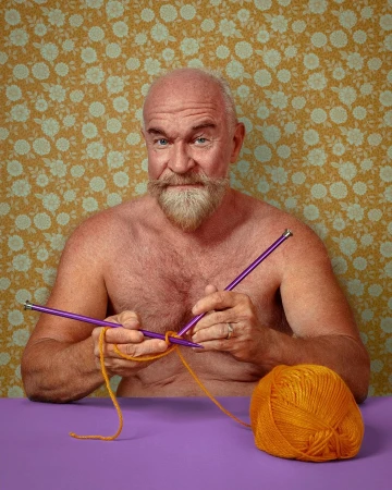 Middle-aged man with beard and bare chest knitting orange yarn with purple knitting needles indoors against a floral wallpaper background.